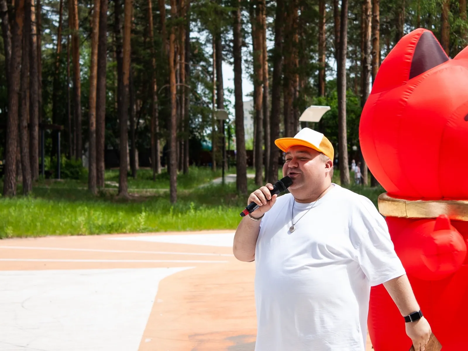 Владимир Шмарёв в белой футболке и кепке с микрофоном на городском празднике в сосновом парке